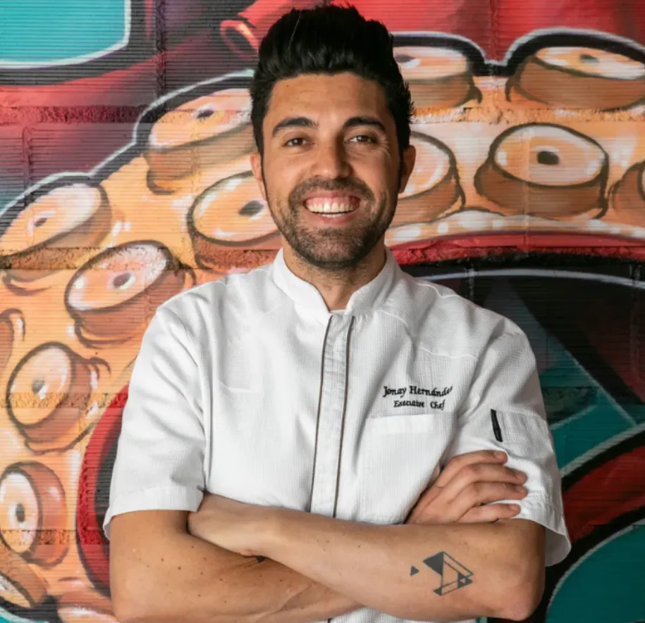 Jonay Hernández, chef ejecutivo, posa frente a un mural de pulpo en su restaurante en Palma. Con chaqueta blanca, tatuaje geométrico y sonrisa confiada, representa la cocina canaria de autor con identidad visual vibrante.