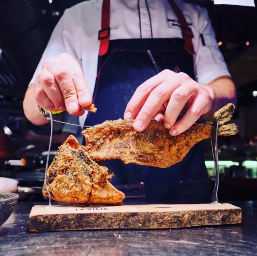 Chef preparando el plato icónico “La Vieja” en cocina profesional. El pescado crujiente se presenta como escultura gastronómica sobre tabla de madera, destacando la técnica y creatividad culinaria.