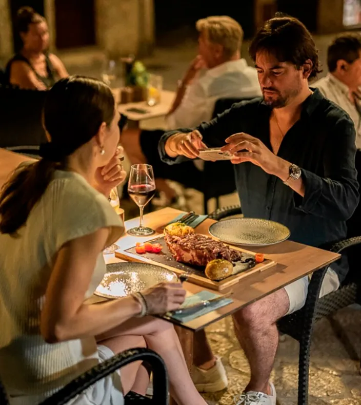 Pareja cenando en La Guarida Palma, restaurante de brasas con fusión japonesa y brasileña. El hombre fotografía un plato principal con carne asada, verduras y vino tinto, mientras la mujer disfruta del ambiente cálido y social del local.