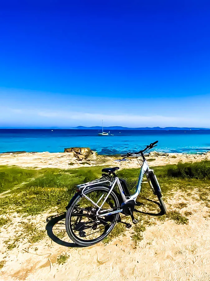 Bicicletas junto al mar turquesa en Formentera con velero al fondo y cielo despejado