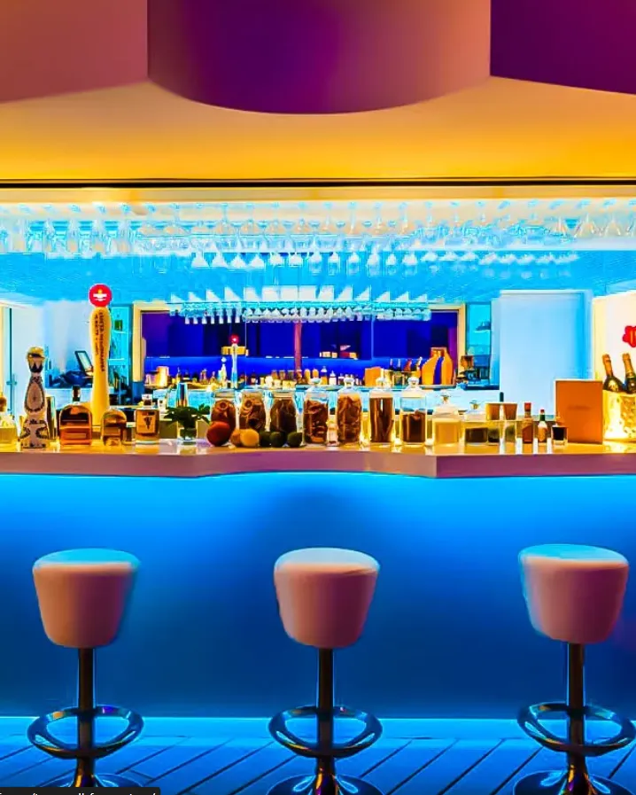 Interior iluminado de The Sky Cocktail Bar Formentera con barra azul, copas colgantes y ambiente moderno